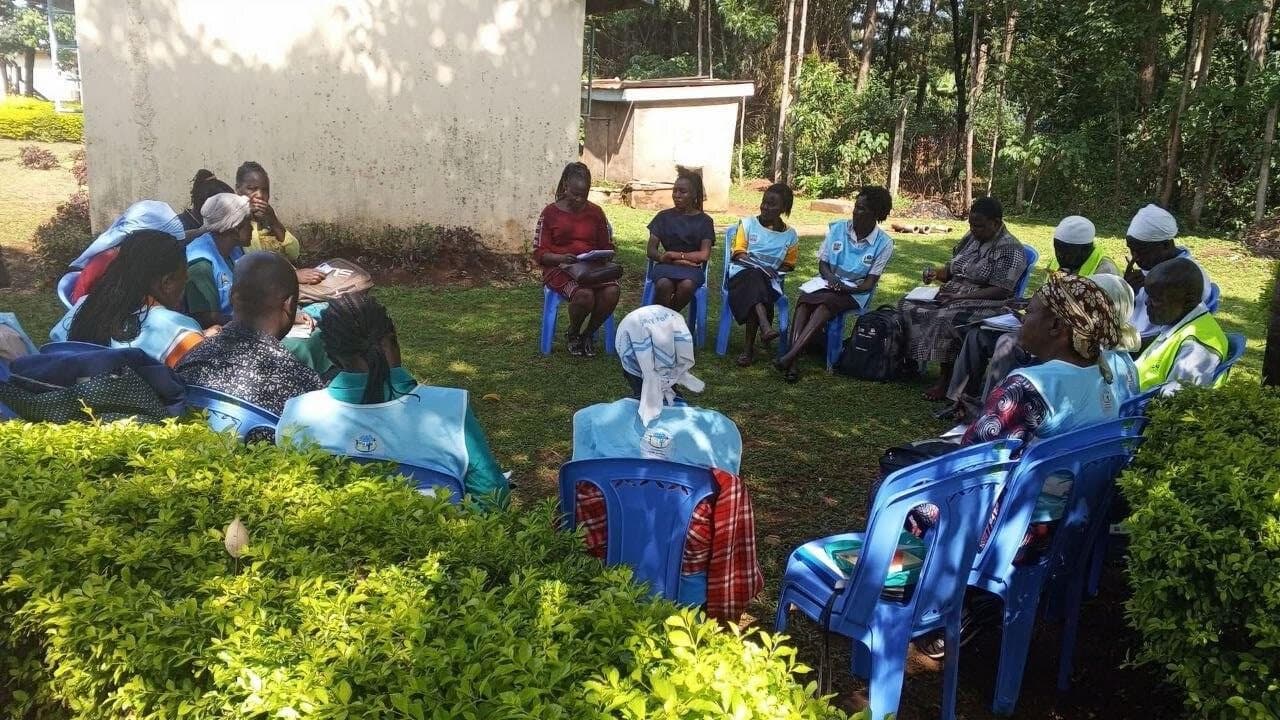 Community health workers seated in a circle on the grass outside a building during a training session.