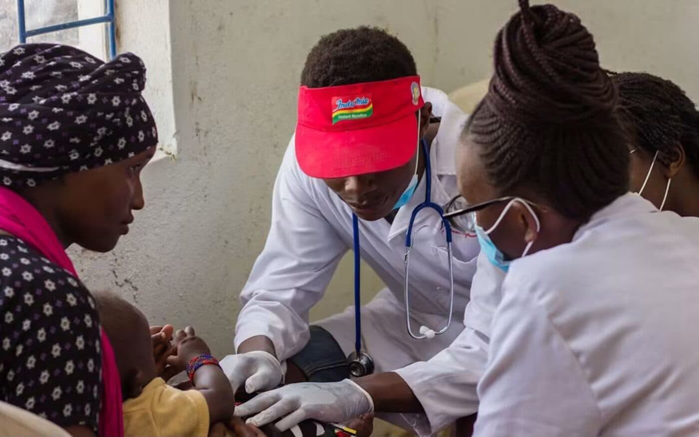 A mother holds her baby steady while two community health workers treat the child at a rural health post.