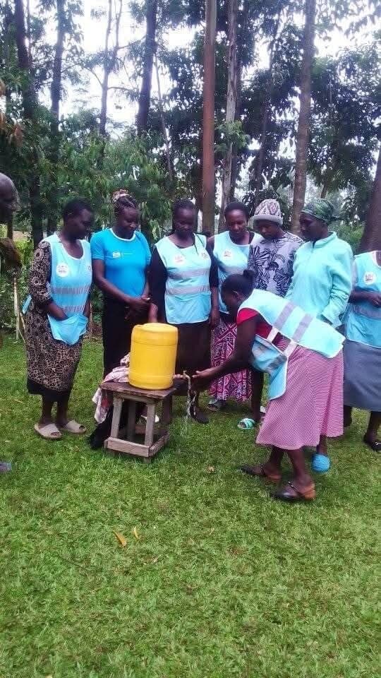 Community health promoters in blue vests gathered around a water container, demonstrating safe water handling to a group of women.